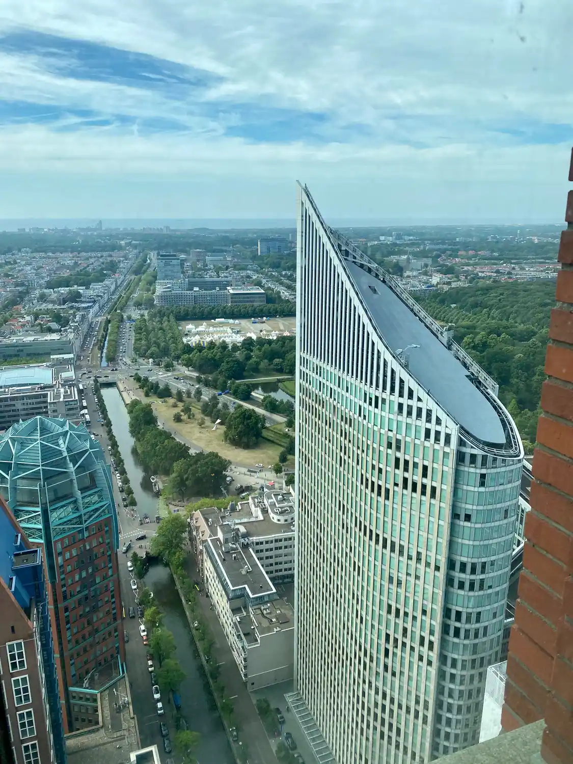 A city seen from a tower, with a green glass slab at the right and roads and trees below.