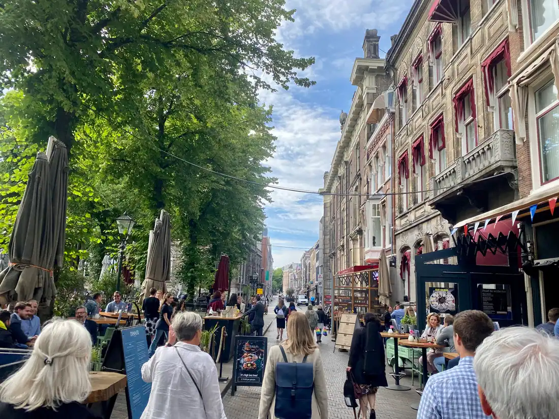 A street of cafe terraces under umbrellas with people seated and walking, a printed menu board and a brick building with red awnings.