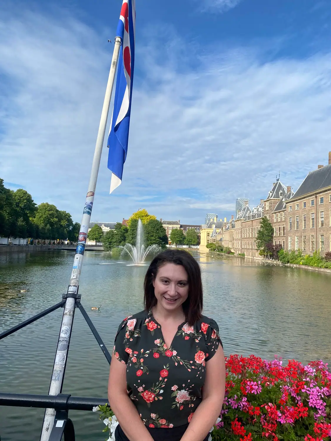 A person standing at a lake with a fountain and a flag on a mast, and a stone building across the water.