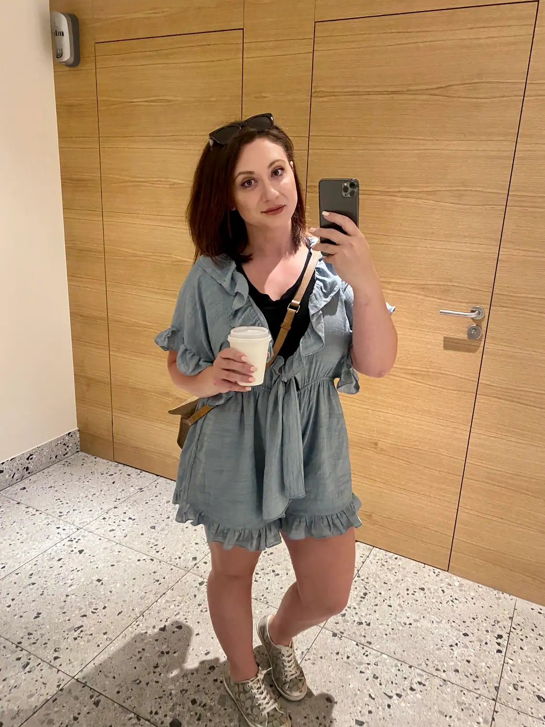A woman taking a picture of herself in a mirror in a timber lined lift lobby, holding a cup.