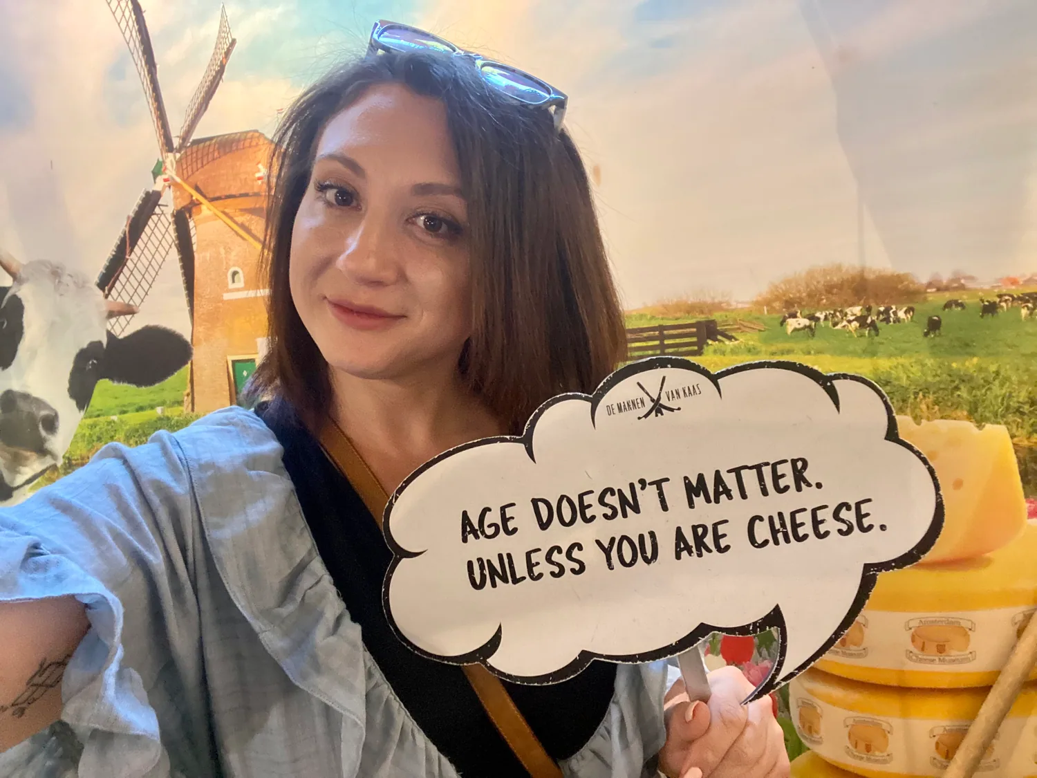 A woman taking a picture of herself against a printed backdrop of a windmill and cows, holding a printed sign, with boxes stacked at the edge.