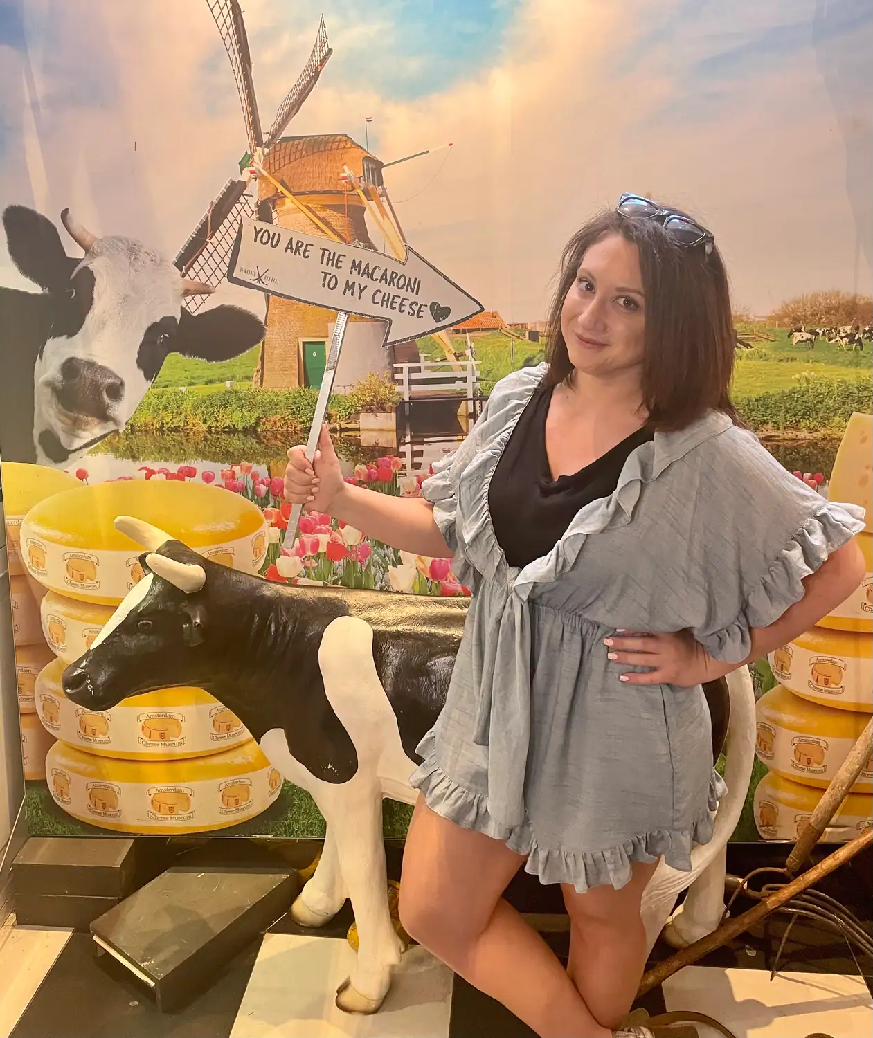 A woman posing against a printed meadow backdrop beside a model cow, holding a printed sign on a staff, with rounds of cheese stacked around.