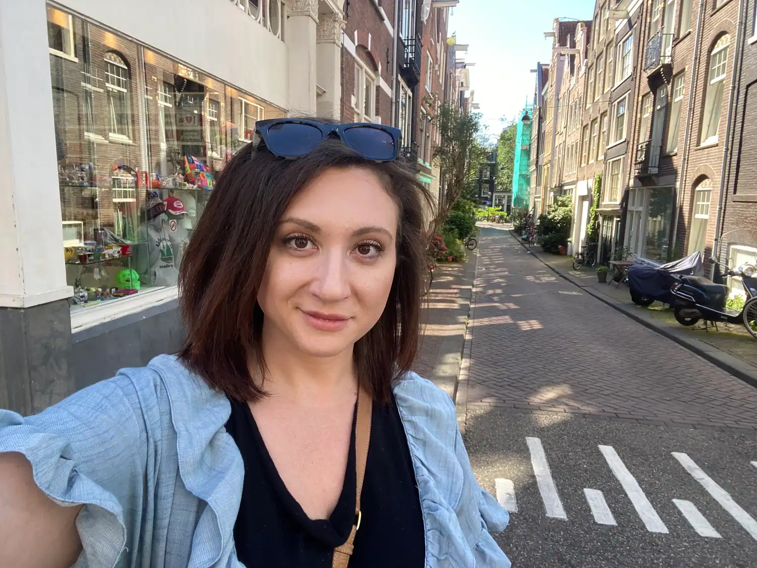 A woman taking a picture of herself in a narrow street with shop windows and parked bikes behind her.