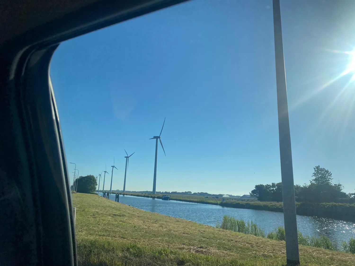 A waterway and embankment with wind turbines seen through a car window, grass and reeds along the bank.