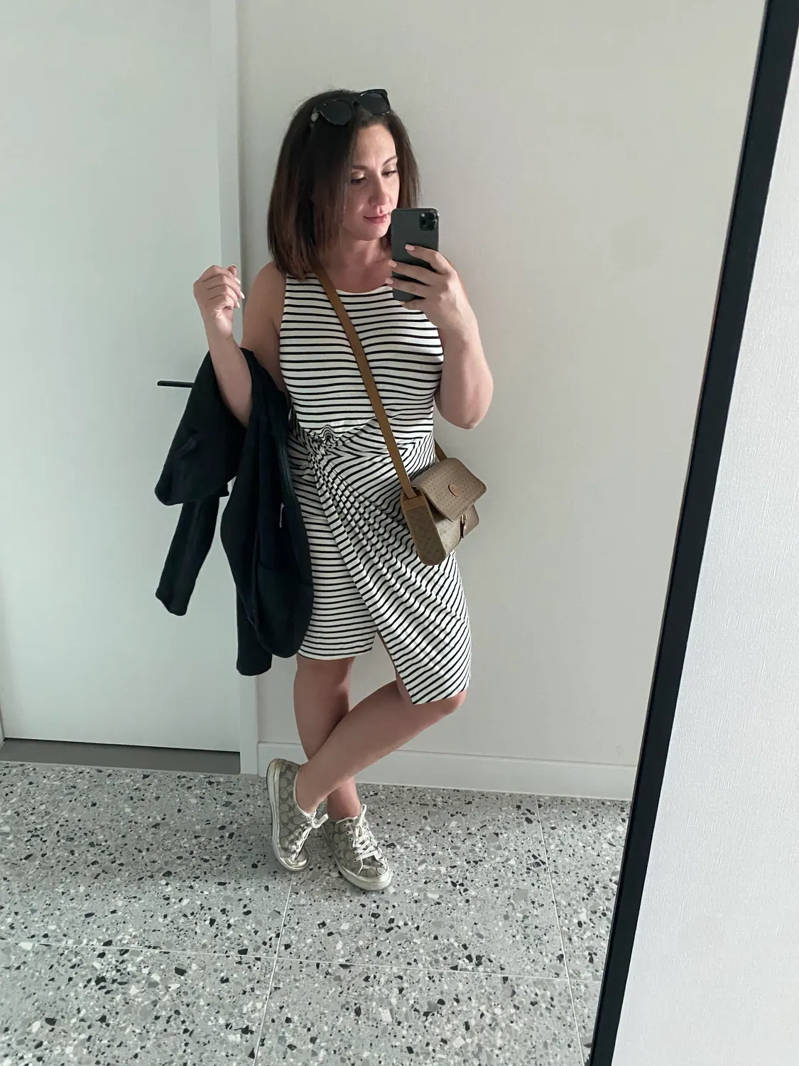A woman taking a picture of herself in a mirror in a plain lobby, a jacket over one arm.