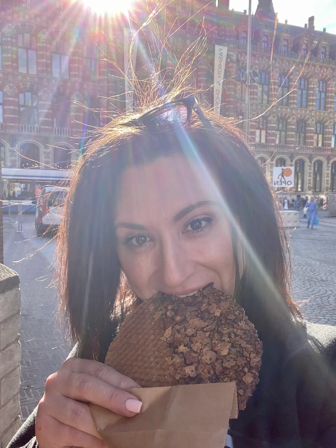 A woman taking a picture of herself in a sunlit square, holding something to eat, with buildings and a few people behind her.