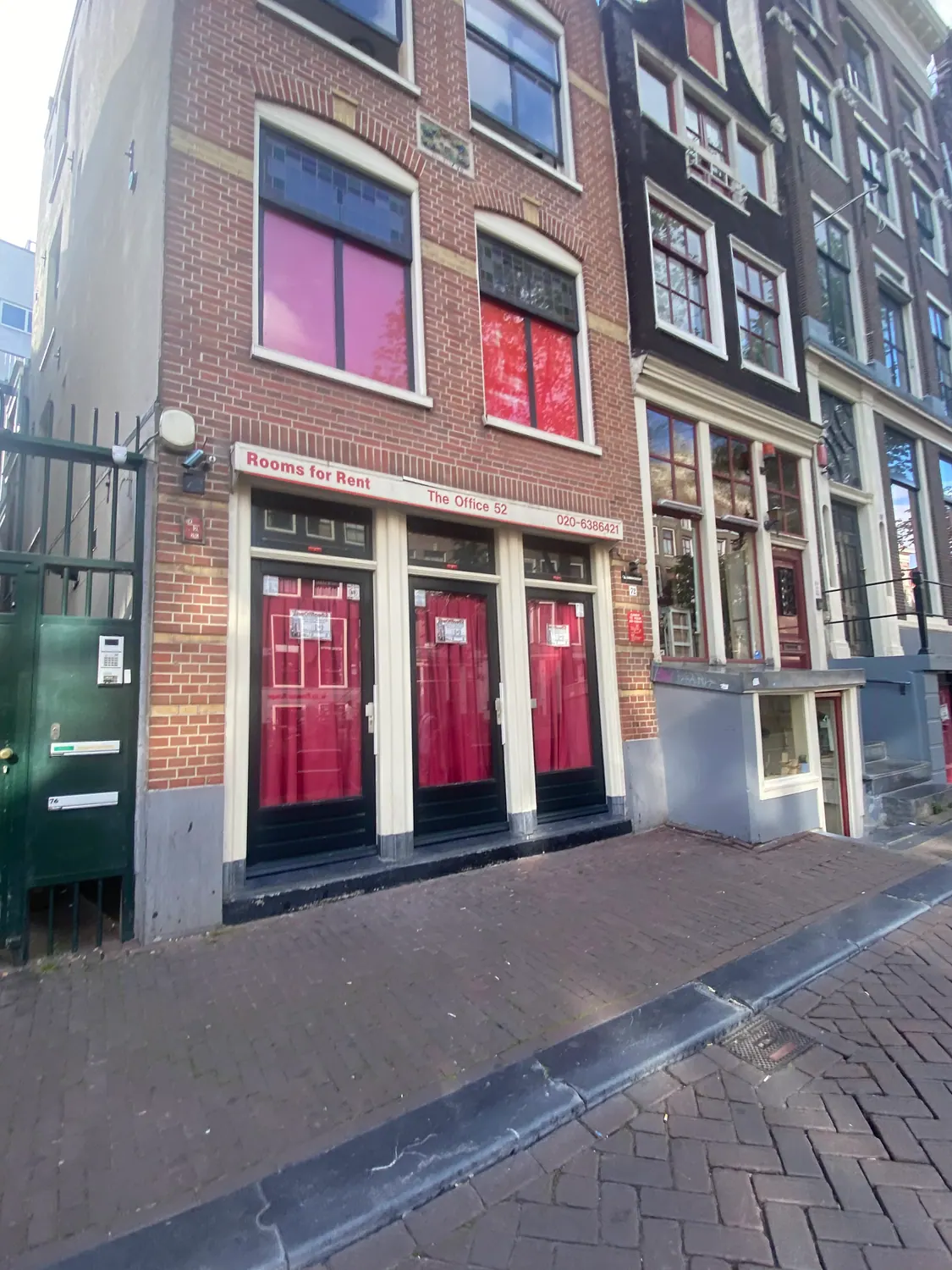 A brick building with red curtained windows and a small sign on its wall, with a gate and paving in front of it.