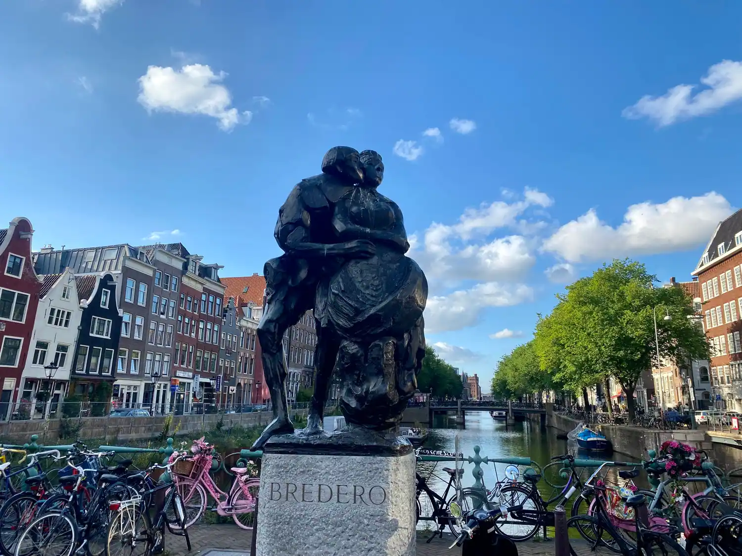 A bronze pair of figures on a stone plinth carrying lettering, with a canal, bridges, trees and parked bicycles behind them.