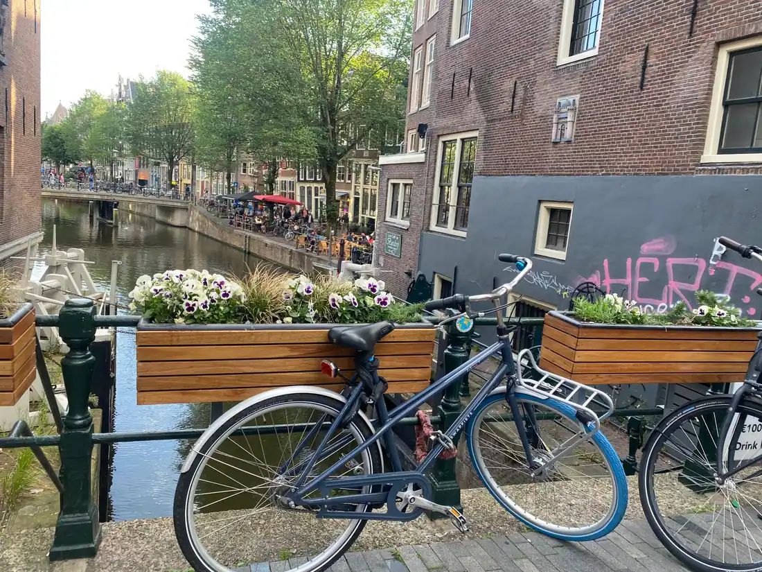 Bicycles and planter boxes along a bridge railing, with a canal below and a wall carrying paint behind.