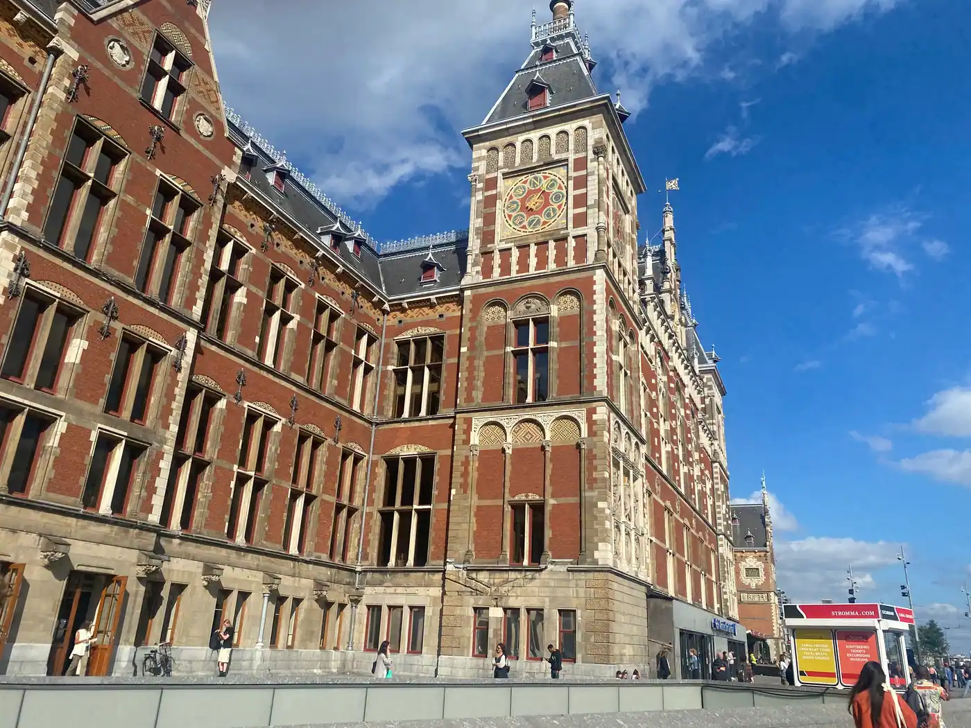 A large brick building with a stone clock tower rising above it, seen from the street, with a few small figures on the pavement below.