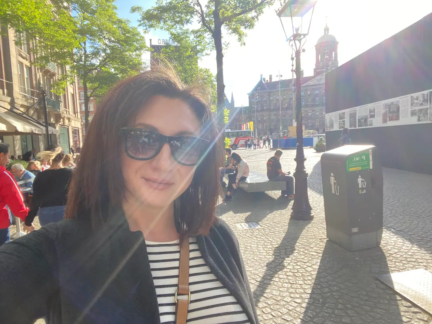 A woman taking a picture of herself in a sunlit square, with buildings and a few people walking behind her.