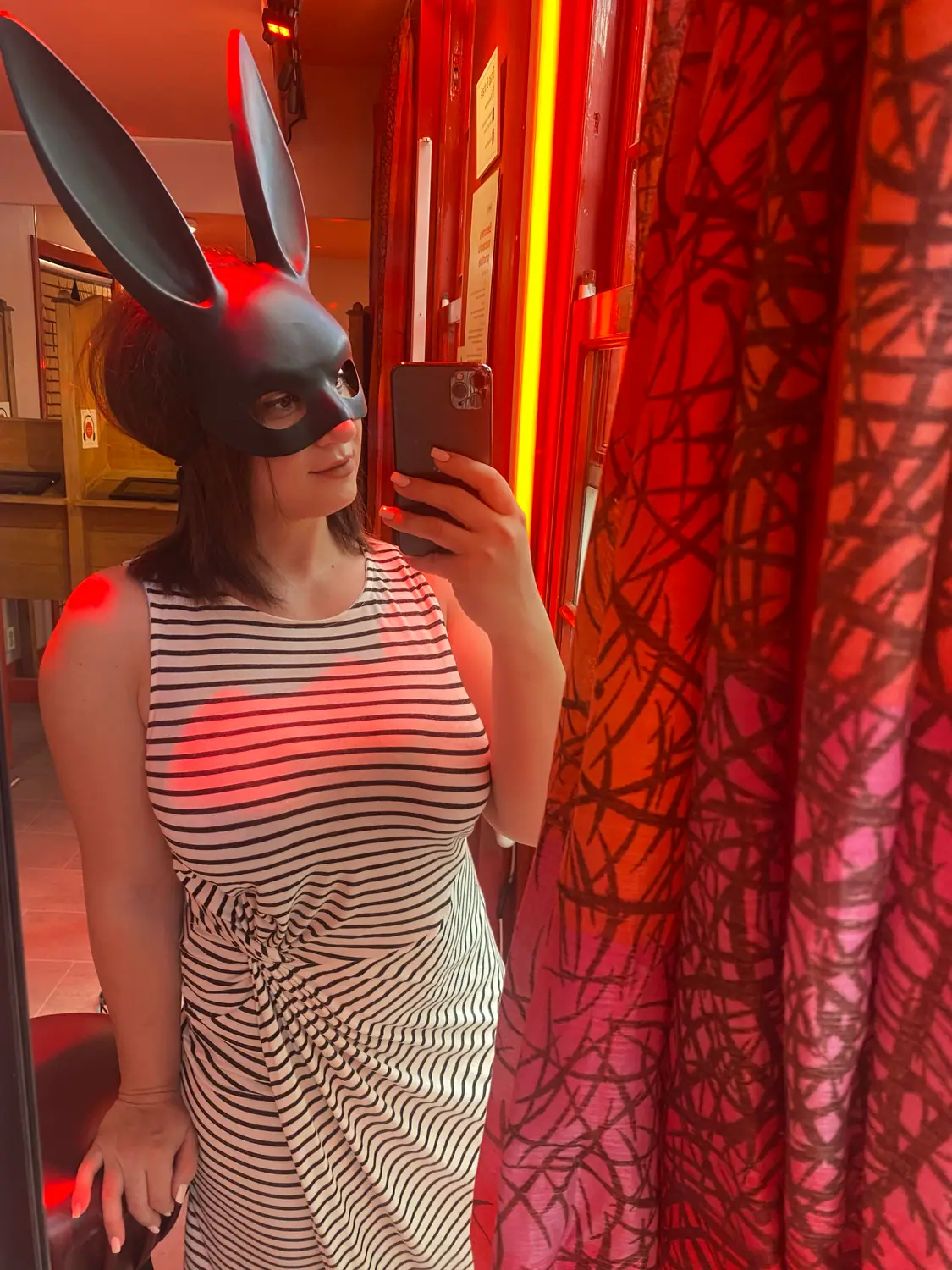 A woman taking a picture of herself in a mirror in a red lit room, wearing a black animal mask, with a patterned curtain behind her.