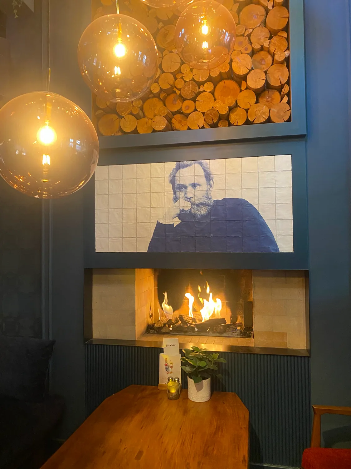 A bar corner with hanging glass lamps, a wall of stacked logs, a fireplace and a framed tiled portrait above a table.
