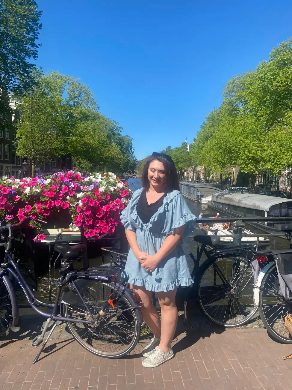 A woman standing among bicycles and flower planters on a canal bridge, with narrow houses along the water behind her.