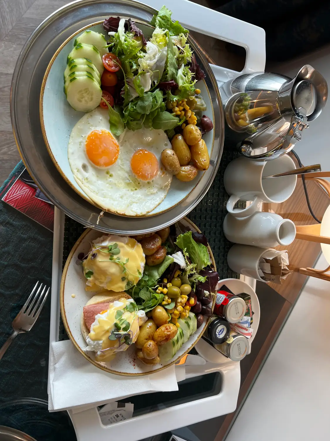 A breakfast tray with a plate of eggs and greens, pastries, a teapot and a glass of juice on a table.