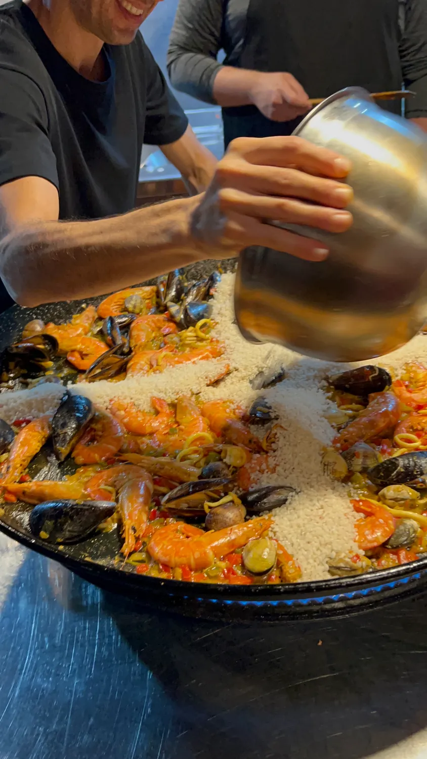 Two hands tipping a jug of broth into a wide shallow pan of rice, an apron behind.
