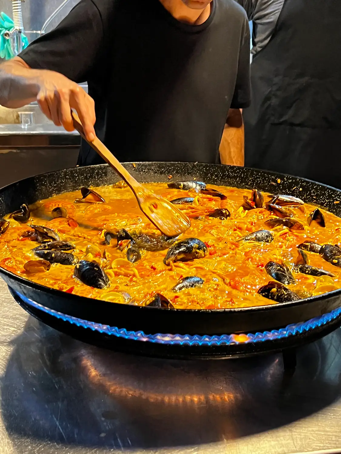 A person stirring a wide pan with a long wooden spoon, seen from the chest down.