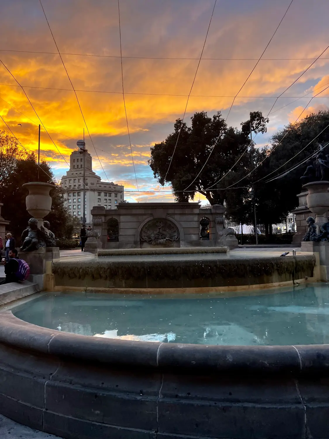 A round fountain basin at dusk with a sculpted figure above it and people sitting on the rim.
