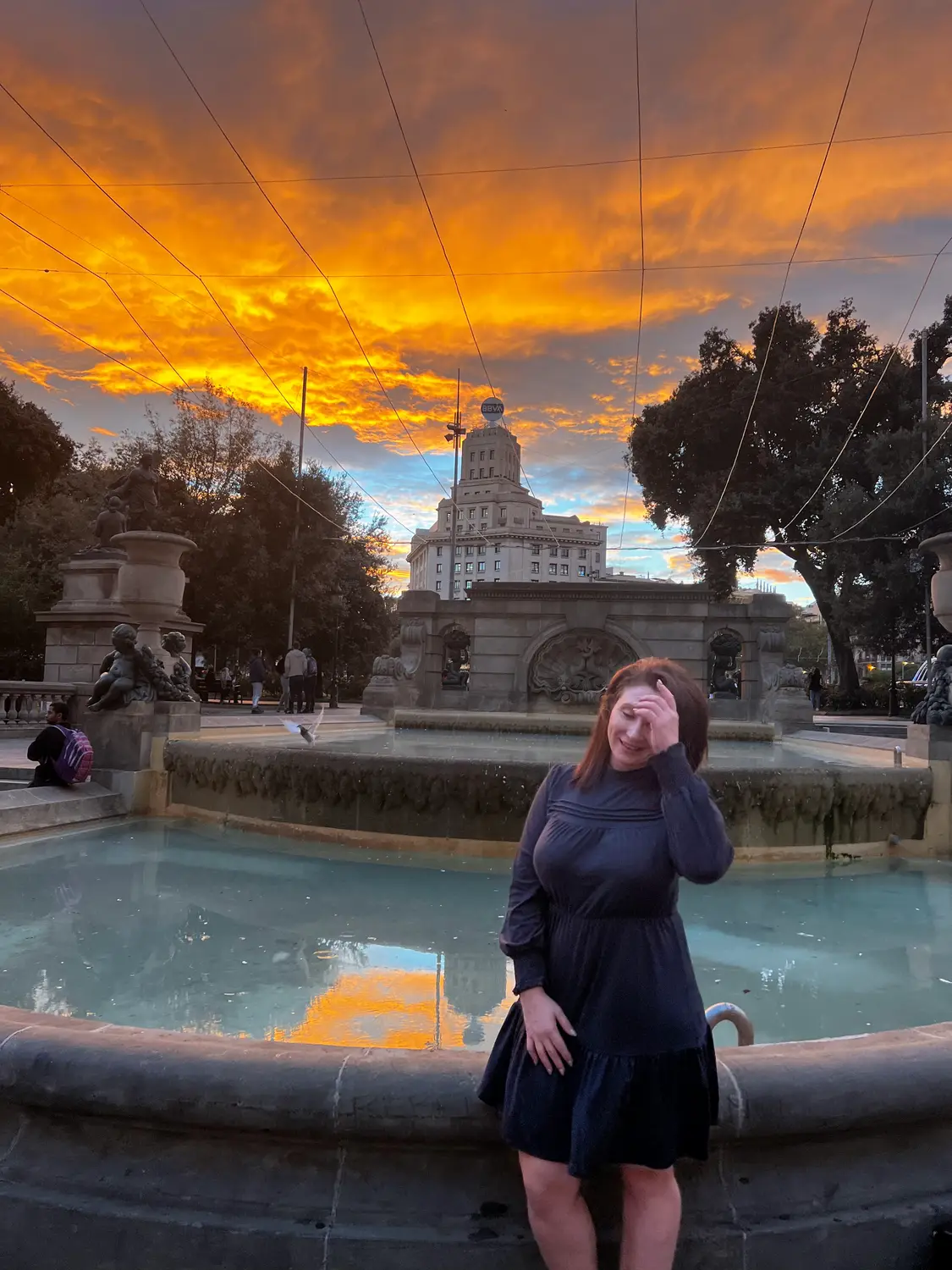 A woman in a dark dress standing by a fountain at dusk, one hand raised, the sunset behind her.