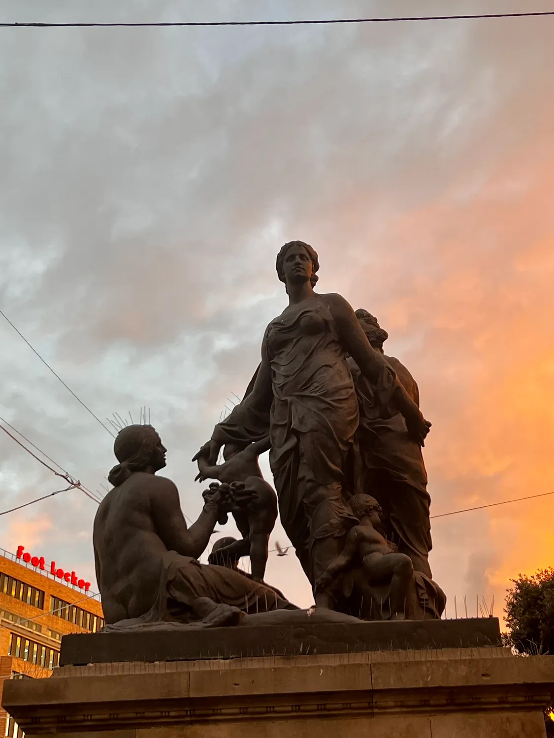 A bronze group of figures on a stone plinth against a sunset sky, shop lights low at the left.
