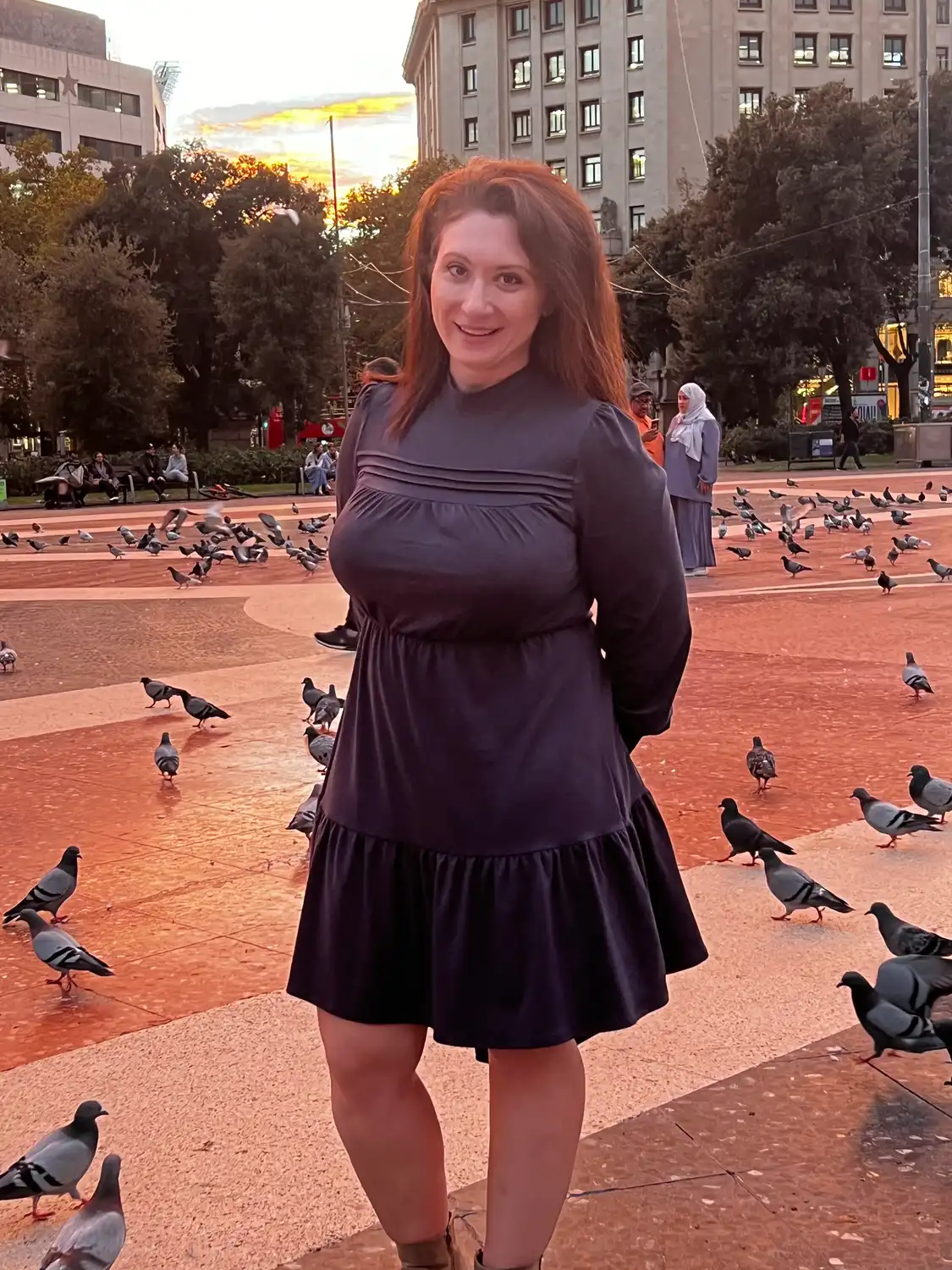 A woman standing in a broad square with pigeons flying past her, palace buildings around.