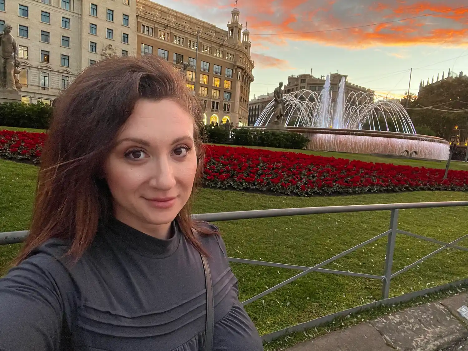 A selfie of a woman at dusk with a civic palace and its fountains behind her.