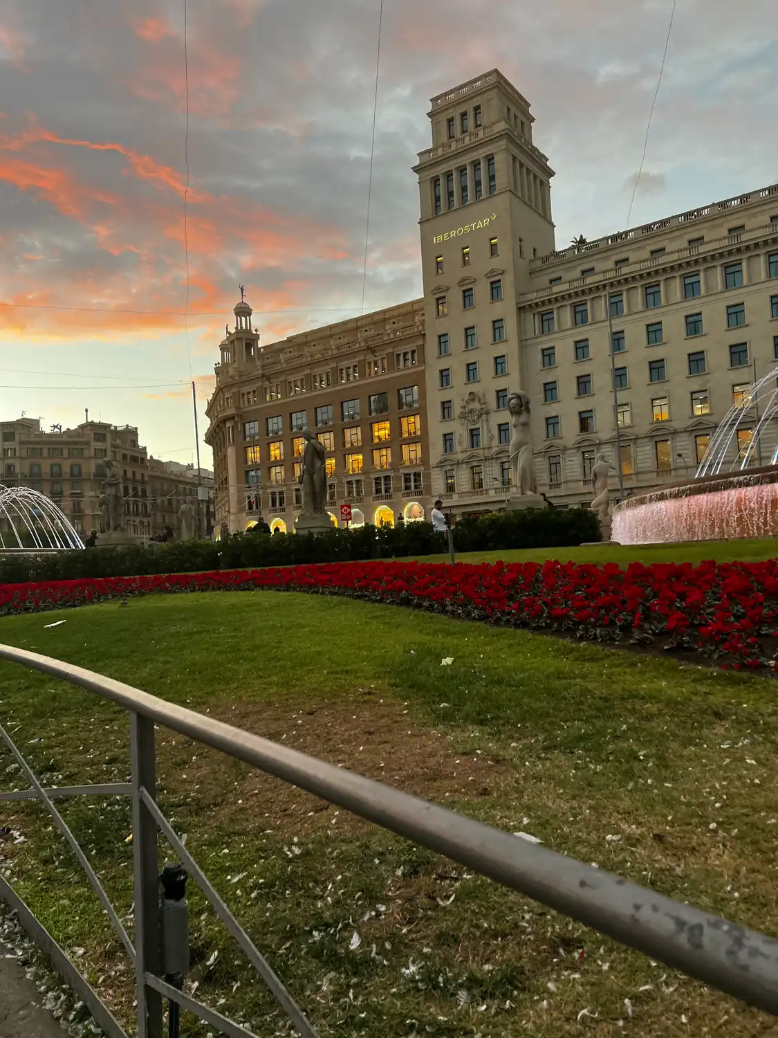 A palace and its gardens seen from a path: clipped hedge, railings and beds of red flowers under cloud.