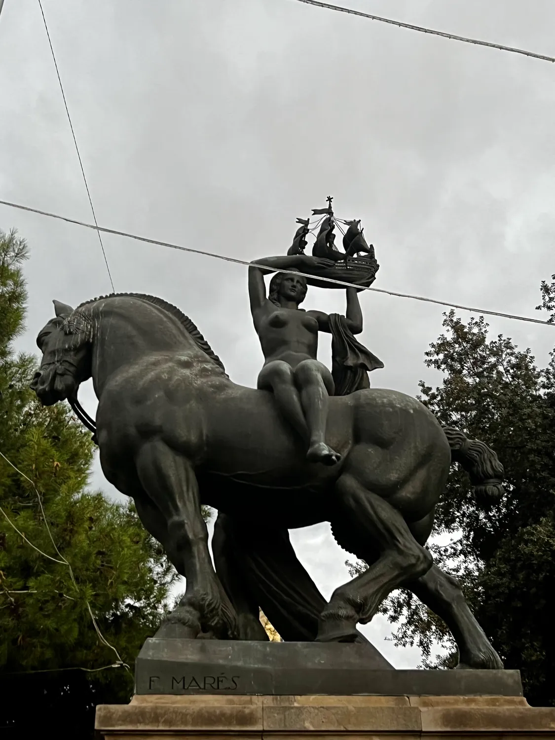 A bronze rider on a horse on a carved stone plinth under a pale sky, trees behind.