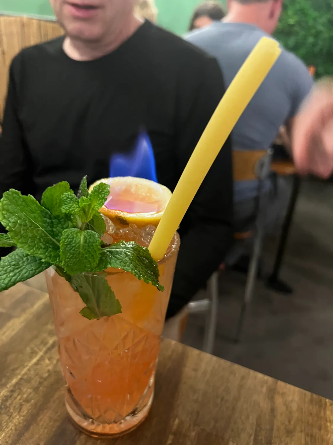 A tall iced drink with mint and a straw on a table, two people seen from behind at a table beyond.