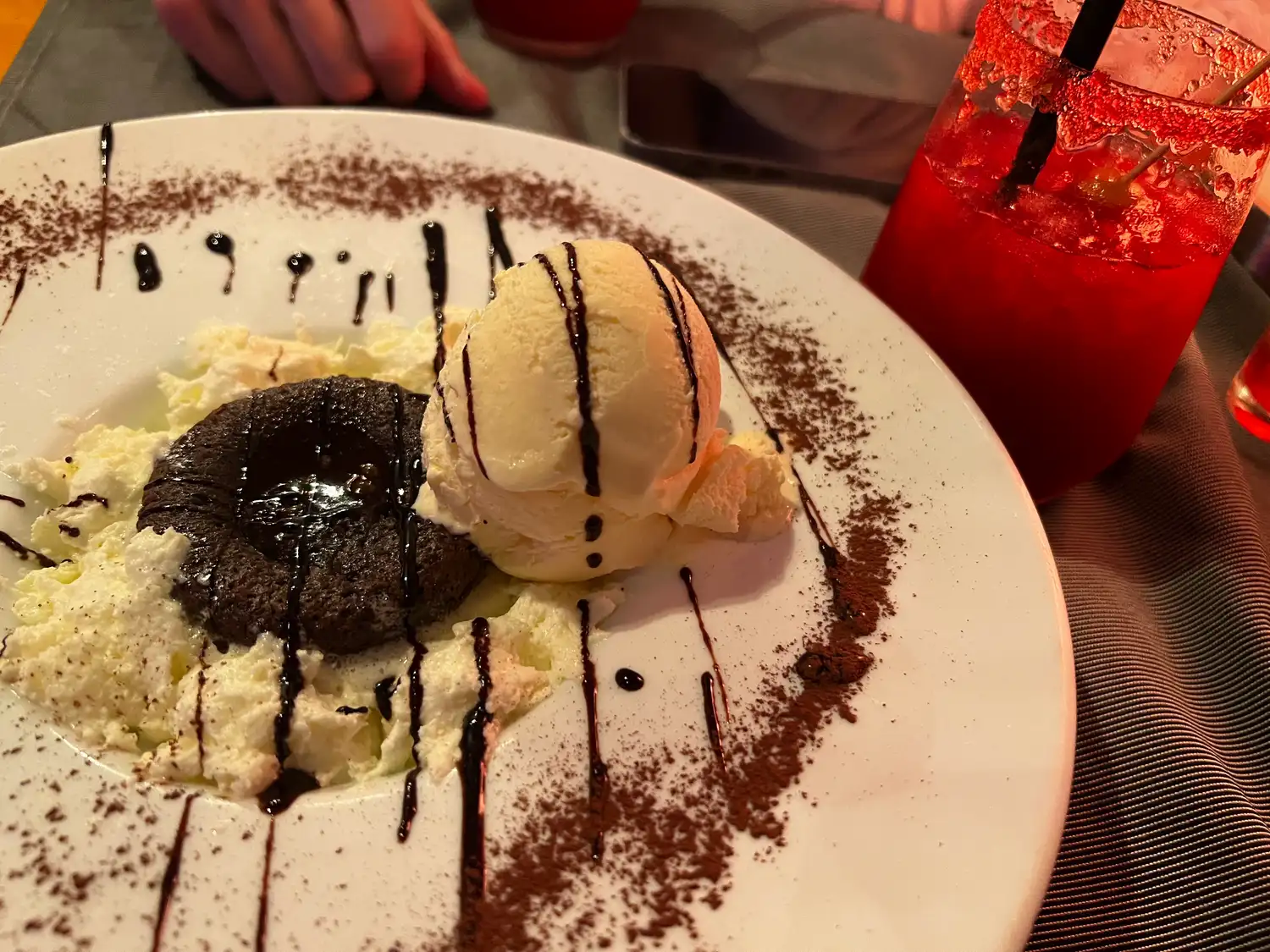 A chocolate dessert with ice cream and sauce on a white plate, a tall red drink behind it.