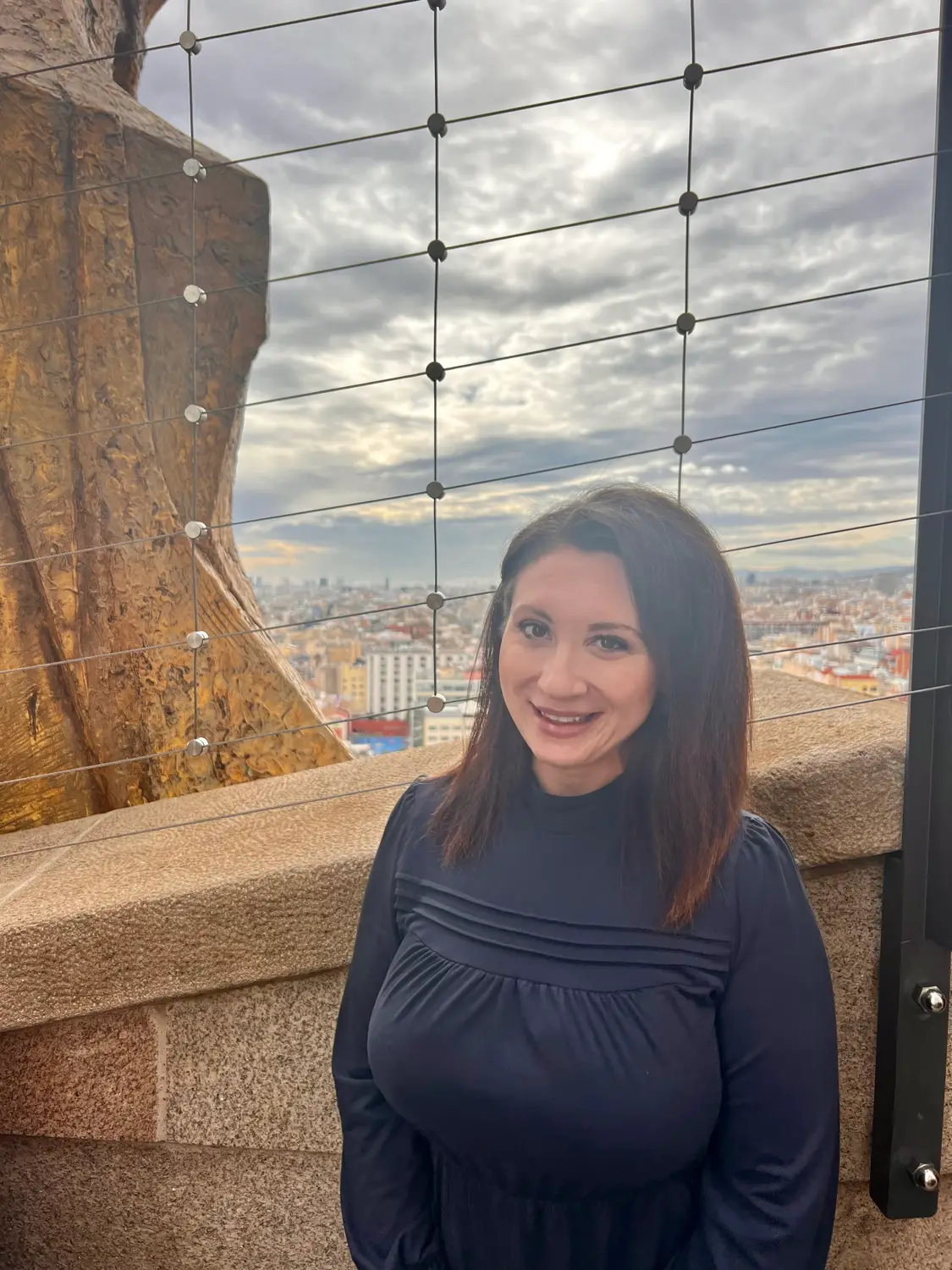 A woman in a dark dress standing at a curved mosaic bench, smiling at the camera, the city far below behind her.
