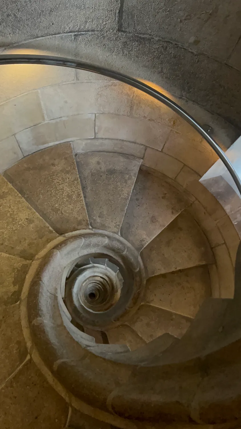 A pale stone spiral staircase seen from below, the steps curling up towards a lit ceiling.