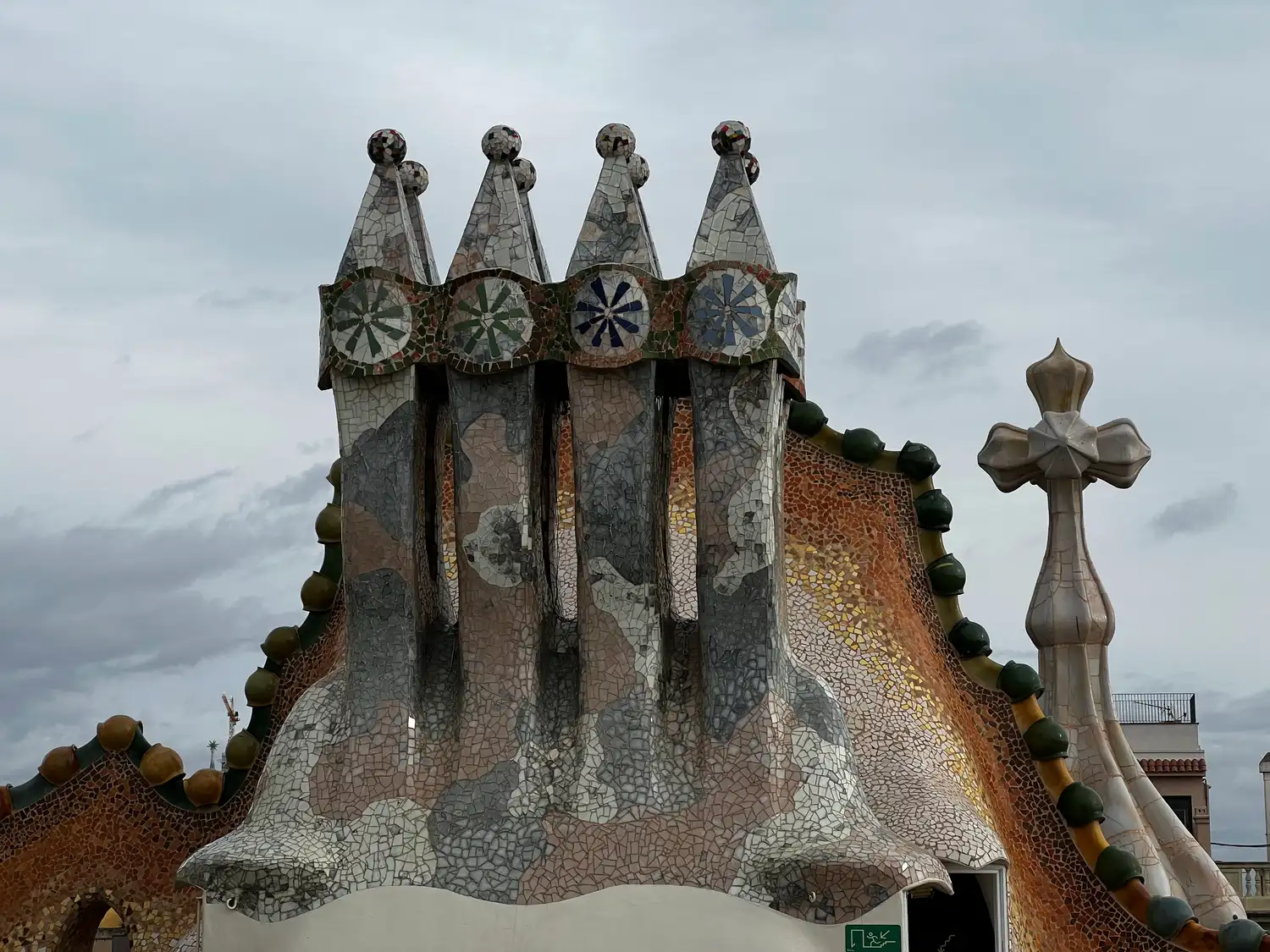Sculpted mosaic-clad roof forms with a small cross on top against grey cloud, the city far behind.