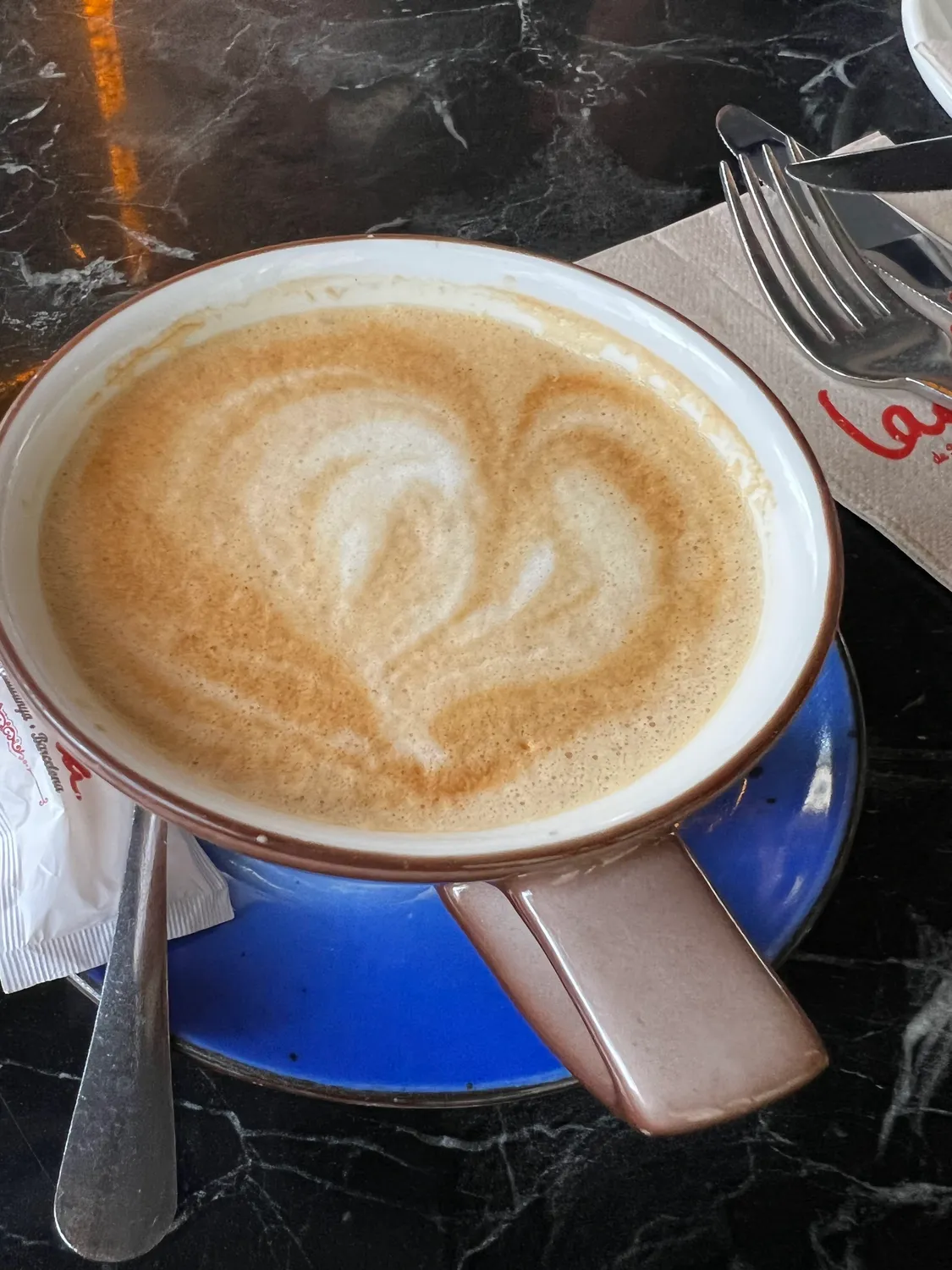 A cup of coffee with a leaf poured into the foam on a saucer, a spoon beside it on the table.