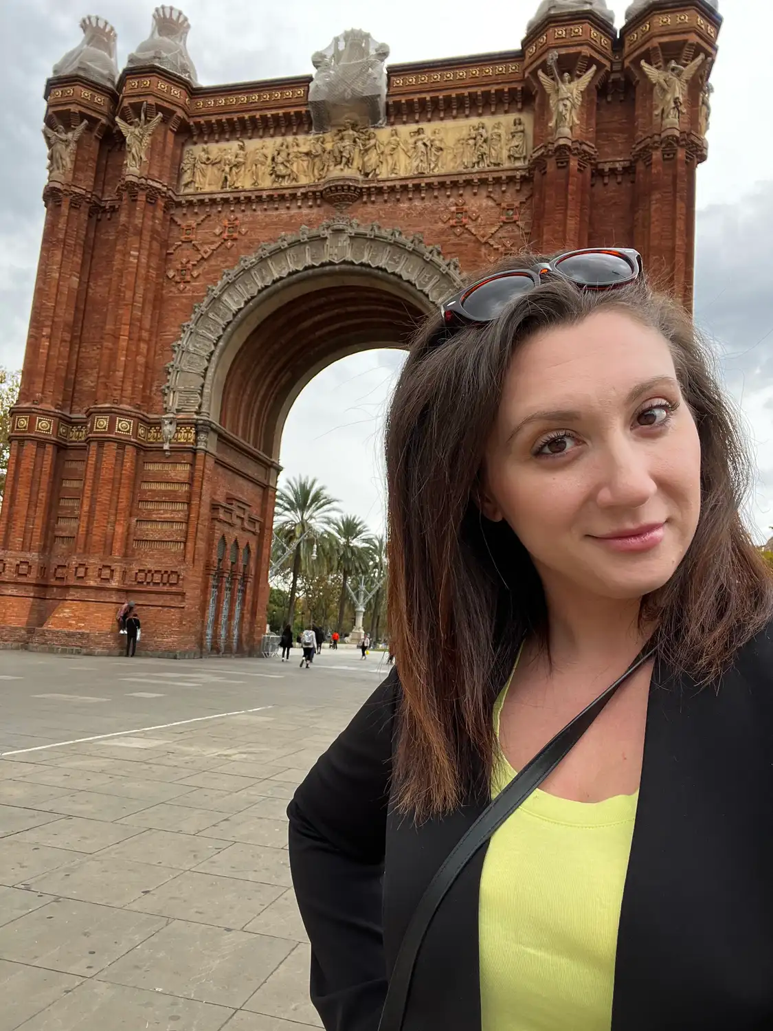 A selfie of a woman in a black jacket and yellow top in front of a brick arch.