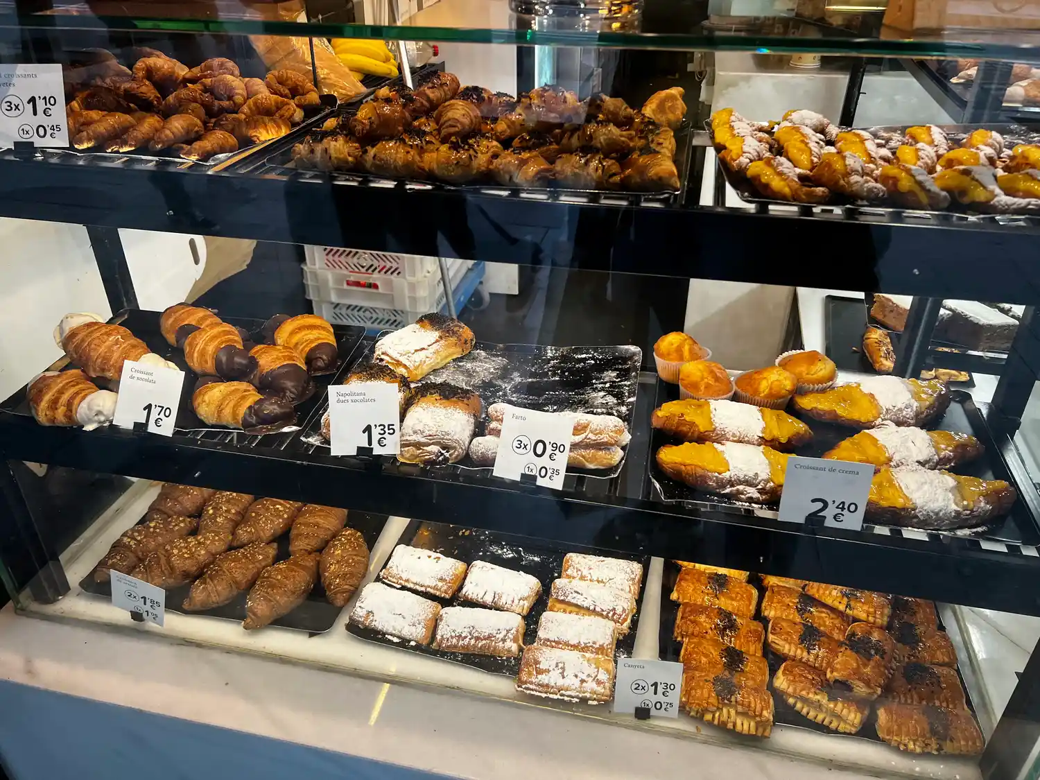 A bakery display case with two shelves of pastries and buns, a small paper card at each tray.
