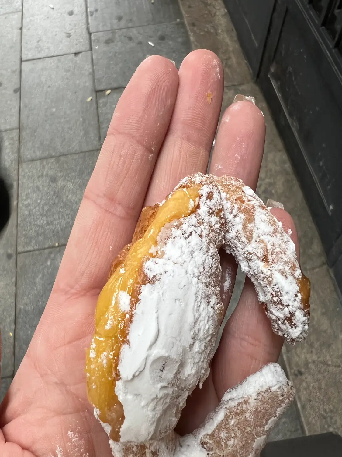 A hand holding a sugar-dusted pastry with a bite taken out of it, paving and a shadow behind.