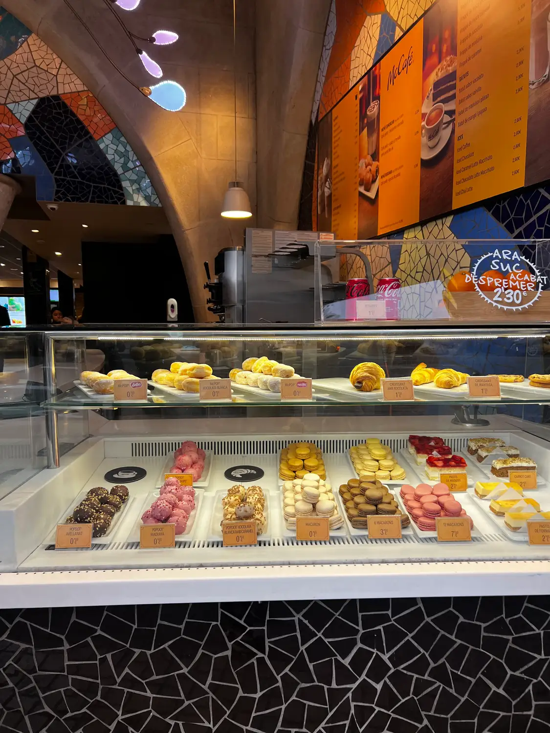 A cafe counter with pastries and small cakes behind glass, a tiled floor and lamps above.