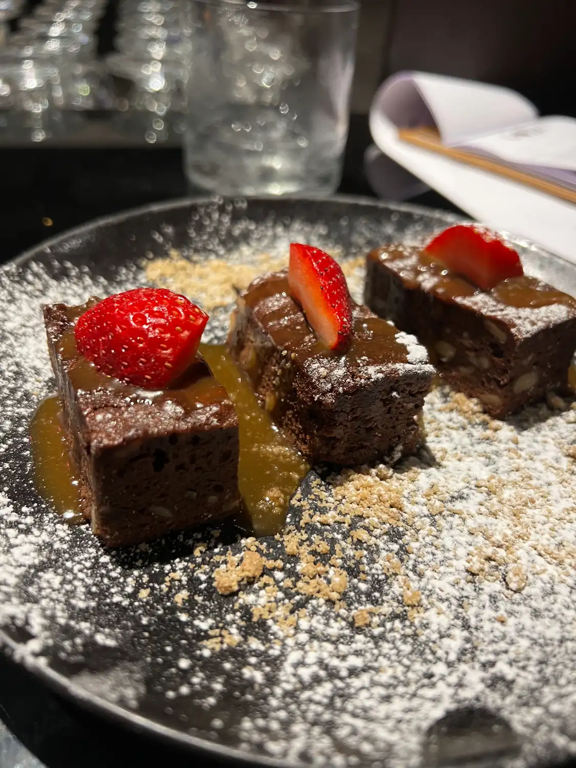 Chocolate squares on a long plate with strawberries and dusted sugar, glassware behind them.