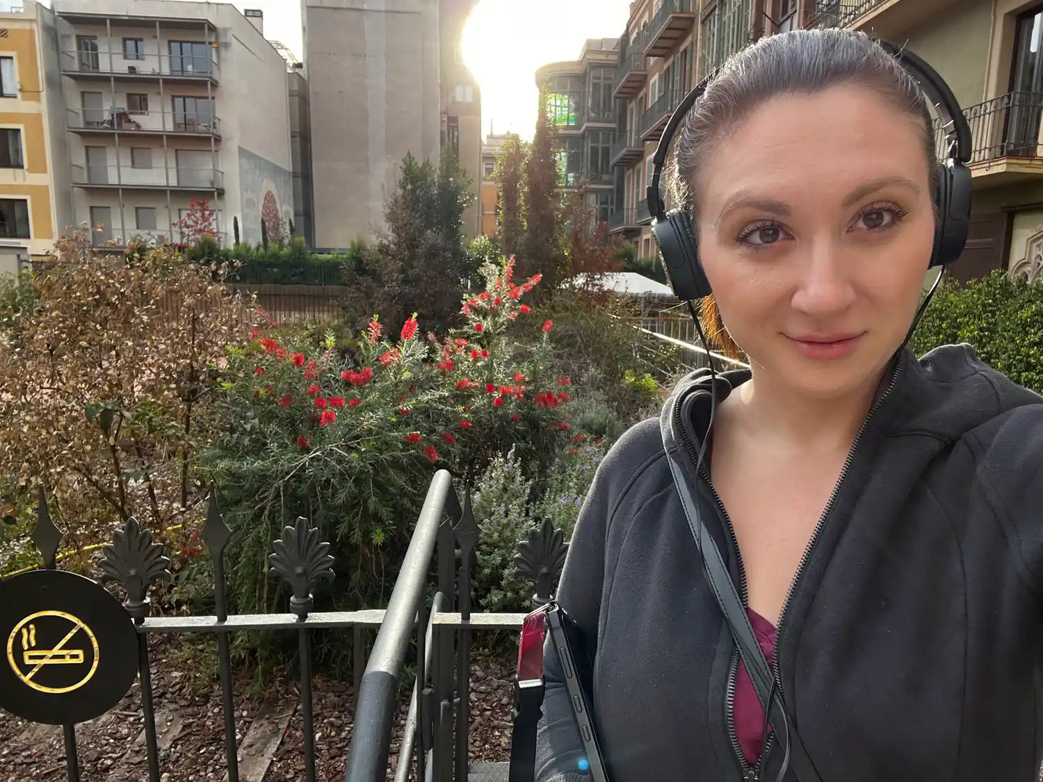 A selfie of a woman wearing headphones in a park, shrubs and a building behind her.