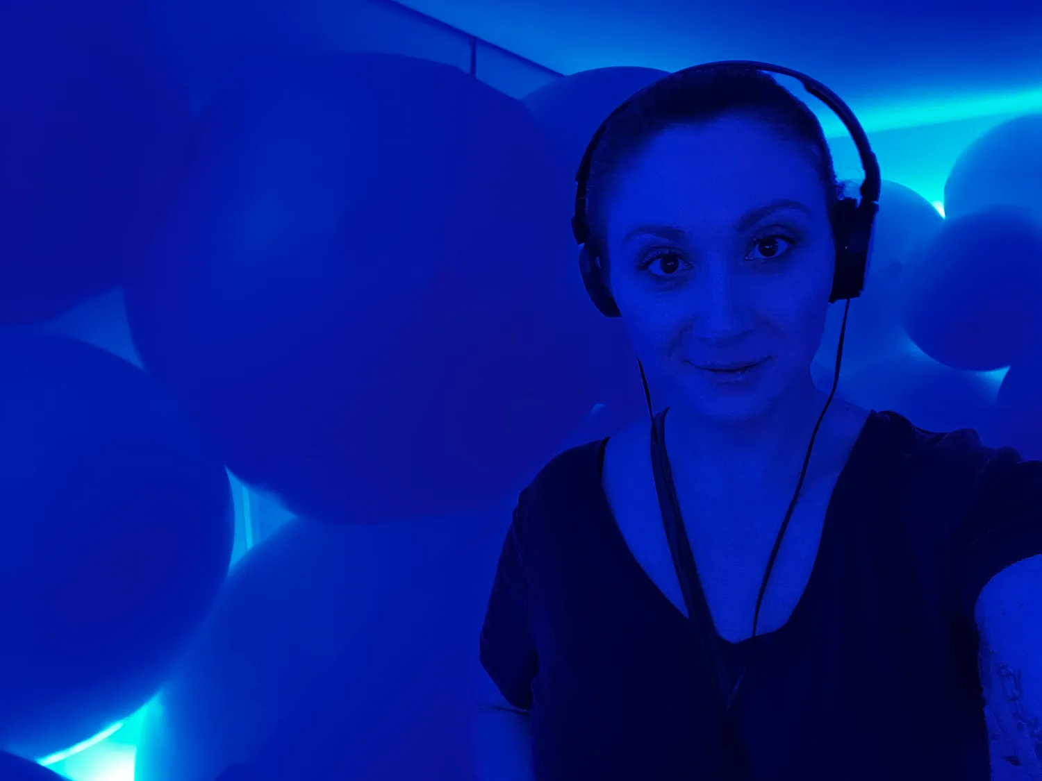 A woman lit only in blue, wearing headphones, looking at the camera in a dark room.