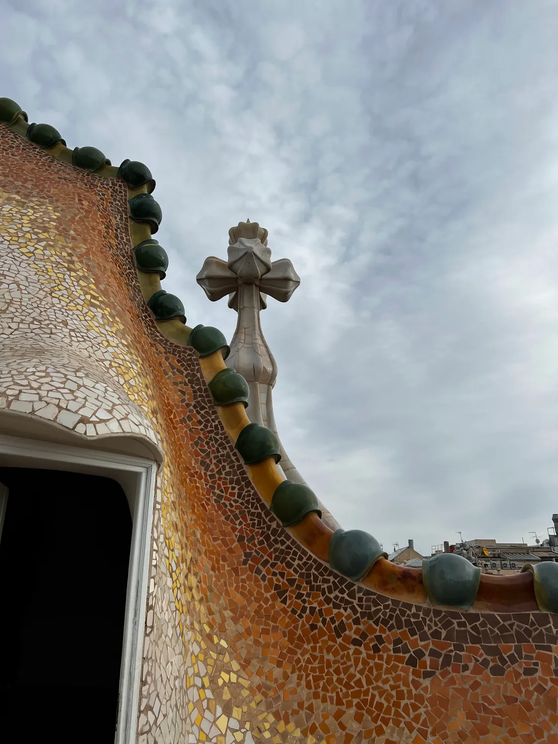 A curved mosaic wall with a cross and small mosaic balls along its edge, sky behind.
