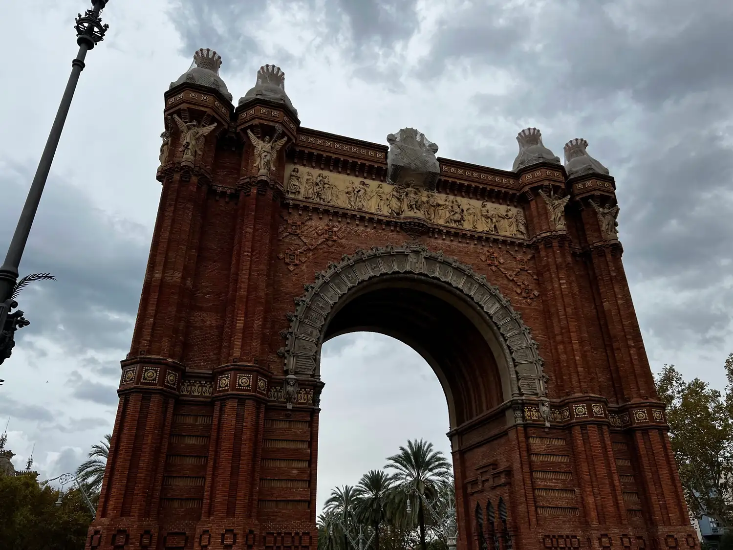 A brick triumphal arch spanning an avenue, with a street lamp and palm trees at either side under cloud.