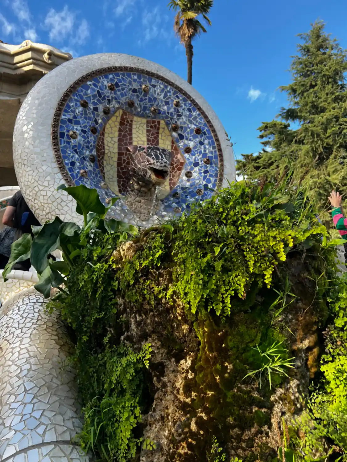 A round mosaic disc set into a wall above green shrubs, with more tilework below it.