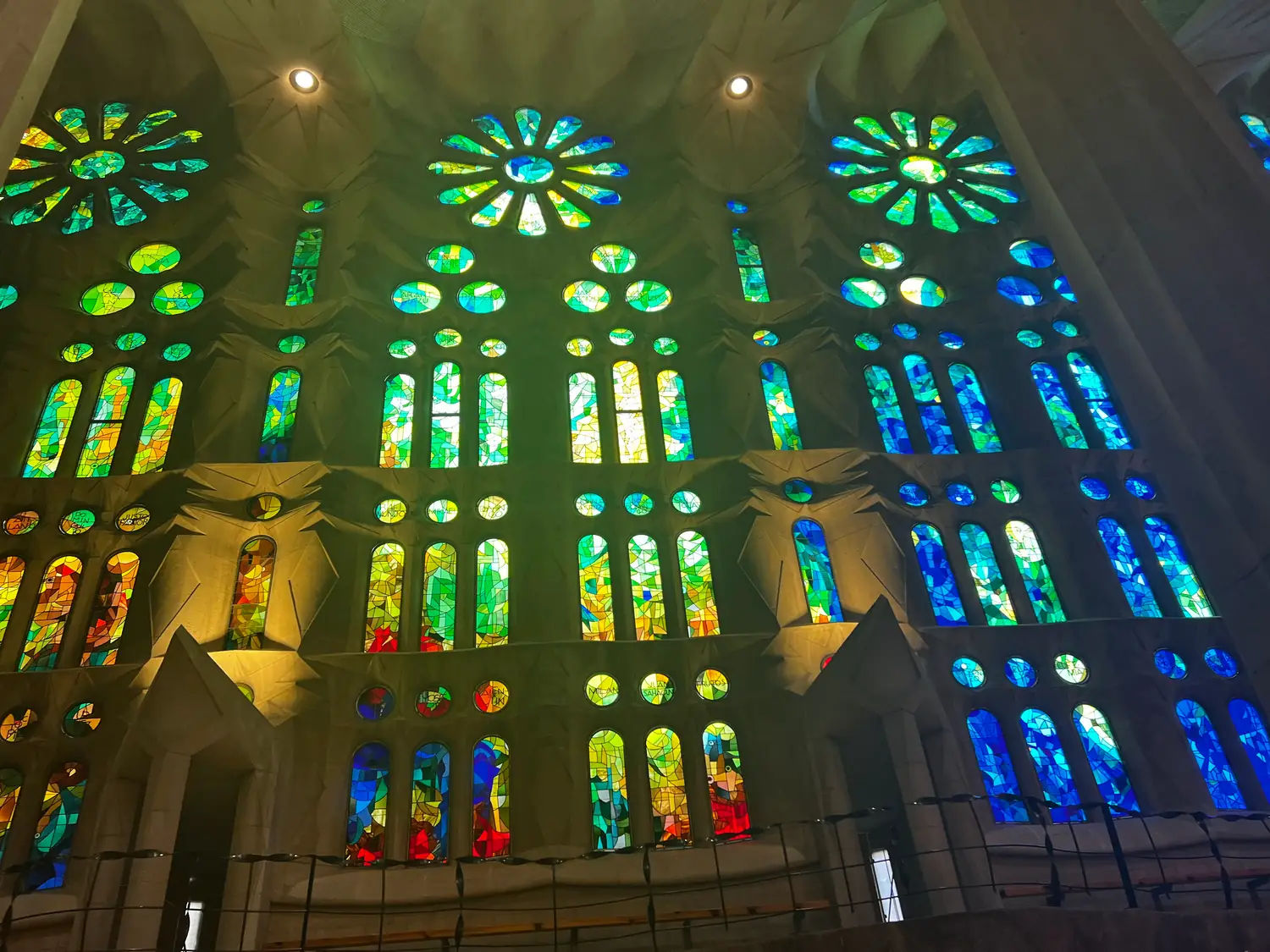 Stained glass windows in blue and green high in a stone wall, with an iron balcony rail in front of them.