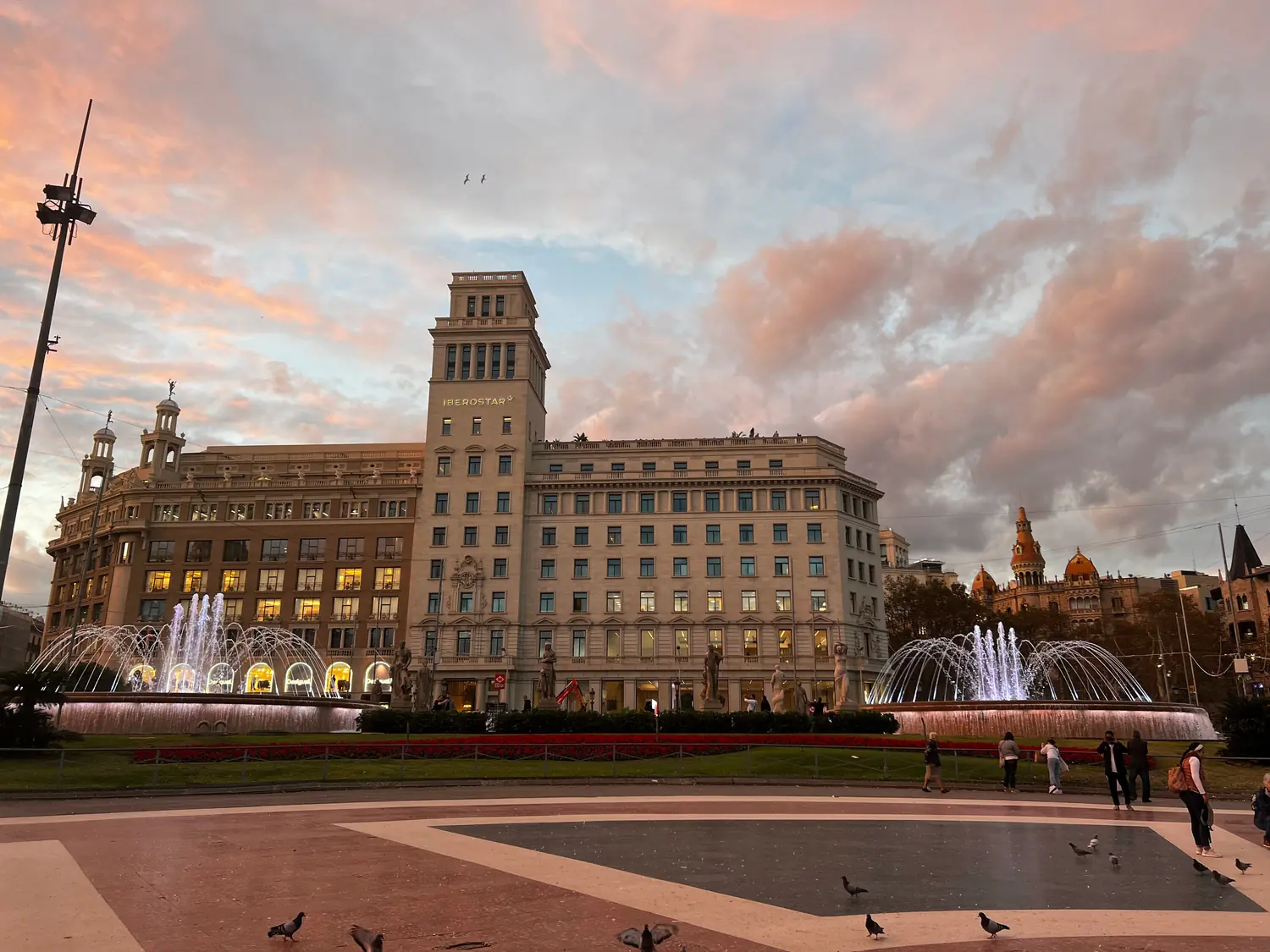 A long civic building with towers and a dome seen across a wide fountain basin at dusk, lamps lit under pink cloud.