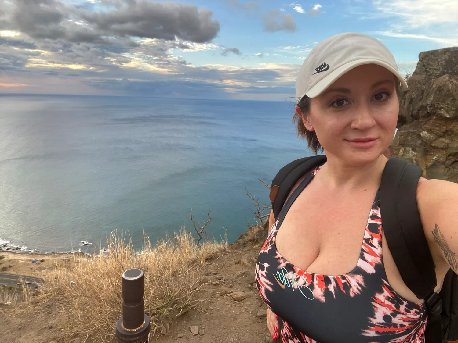 A woman in a white cap and a printed top taking a picture of herself on a ridge, with a metal post beside her and the sea and the coast below.