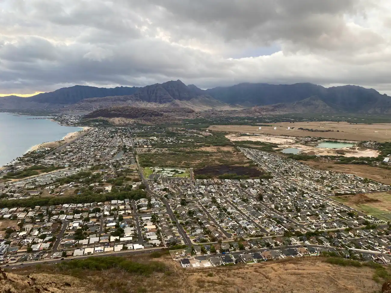 A wide view over a town and its mountain range from a ridge, with a beach, the sea and heavy cloud beyond.