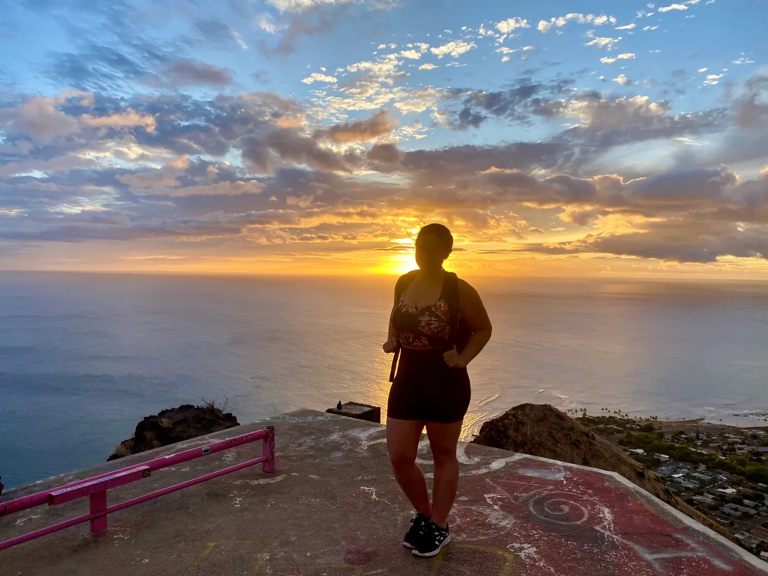 A woman seen from behind in silhouette standing on a concrete lookout platform above the sea, with the sun low ahead of her and a rail at the left.