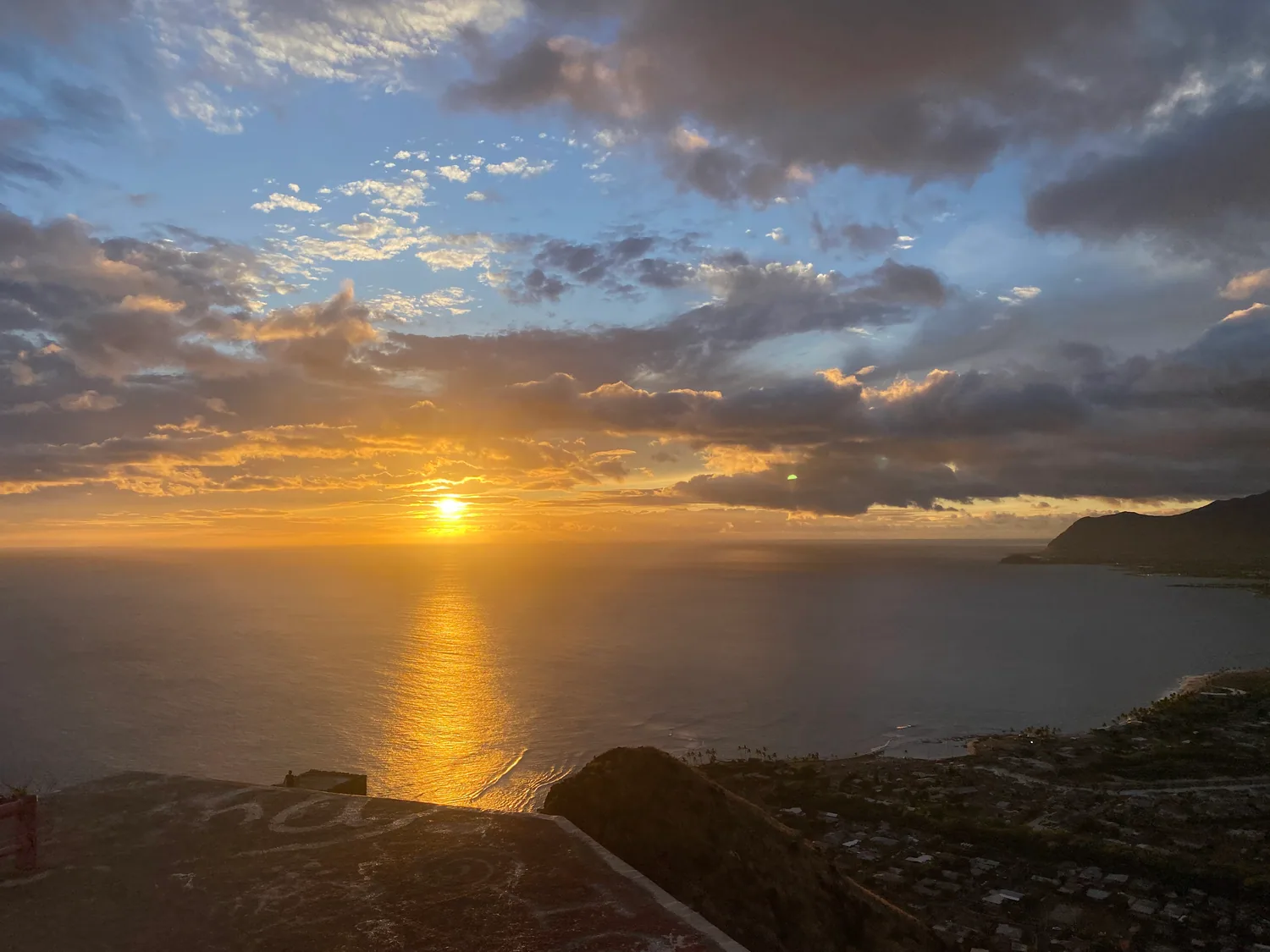 The sun setting into the sea with cloud above it, seen over the concrete of a lookout platform in the foreground, with the town and the coast along the right.